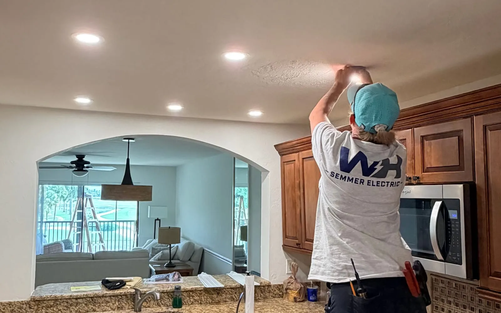 WH Semmer Electric crew member installing recessed lighting in a Fort Myers kitchen ceiling, with a ceiling fan in the room beyond